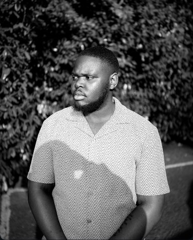 A man with a serious expression stands outdoors against a leafy background.