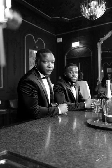 Two men in tuxedos sitting at a bar, with drinks and a glamorous setting in the background.